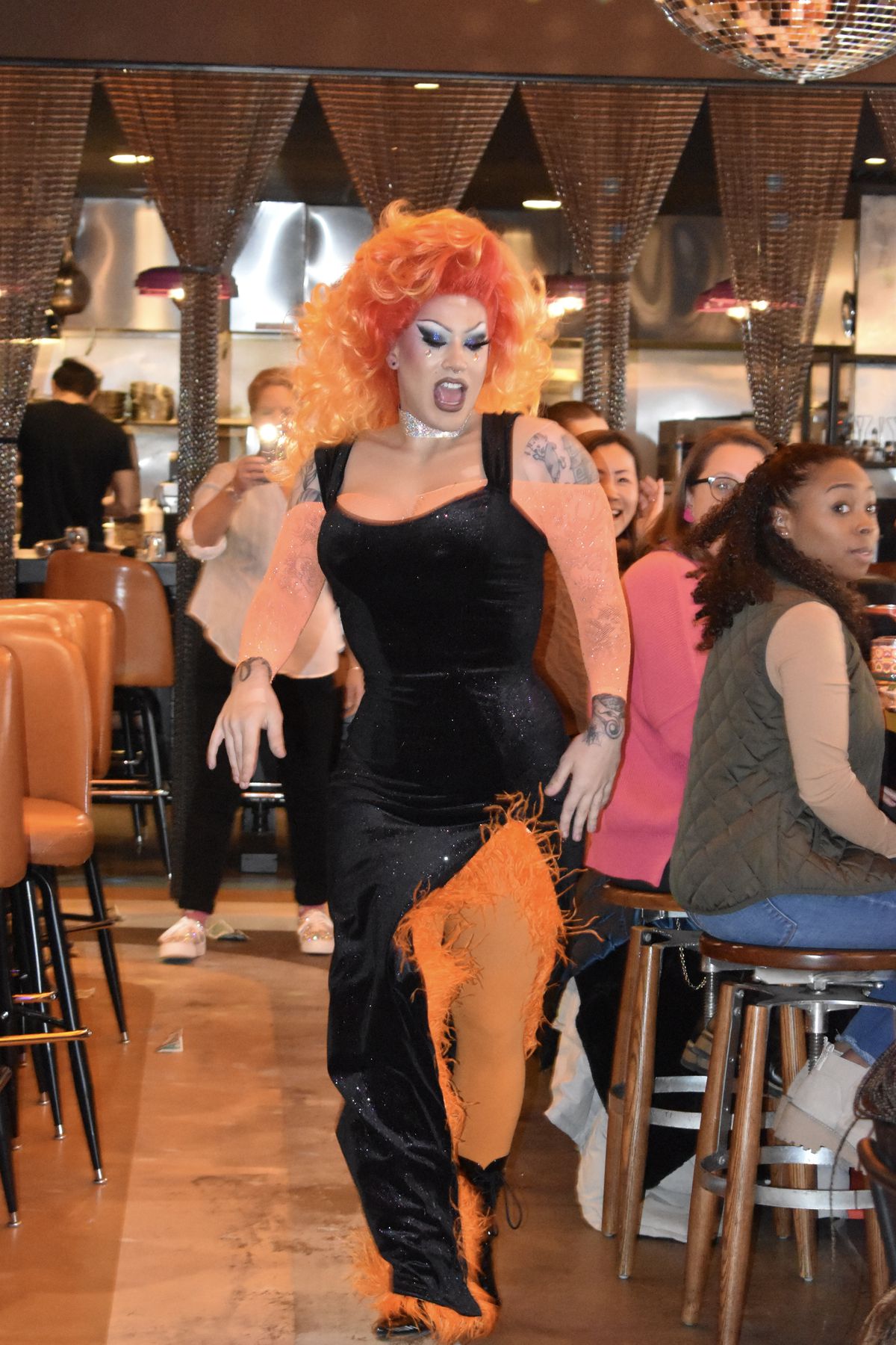A drag performer in a bright orange wig and black and orange dress dances through a dining room.