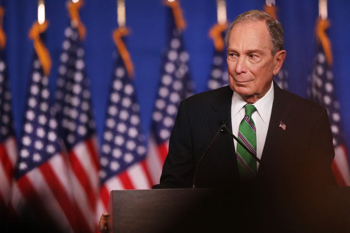 Michael Bloomberg, wearing a green tie, stands at a podium in front of a row of American flags.