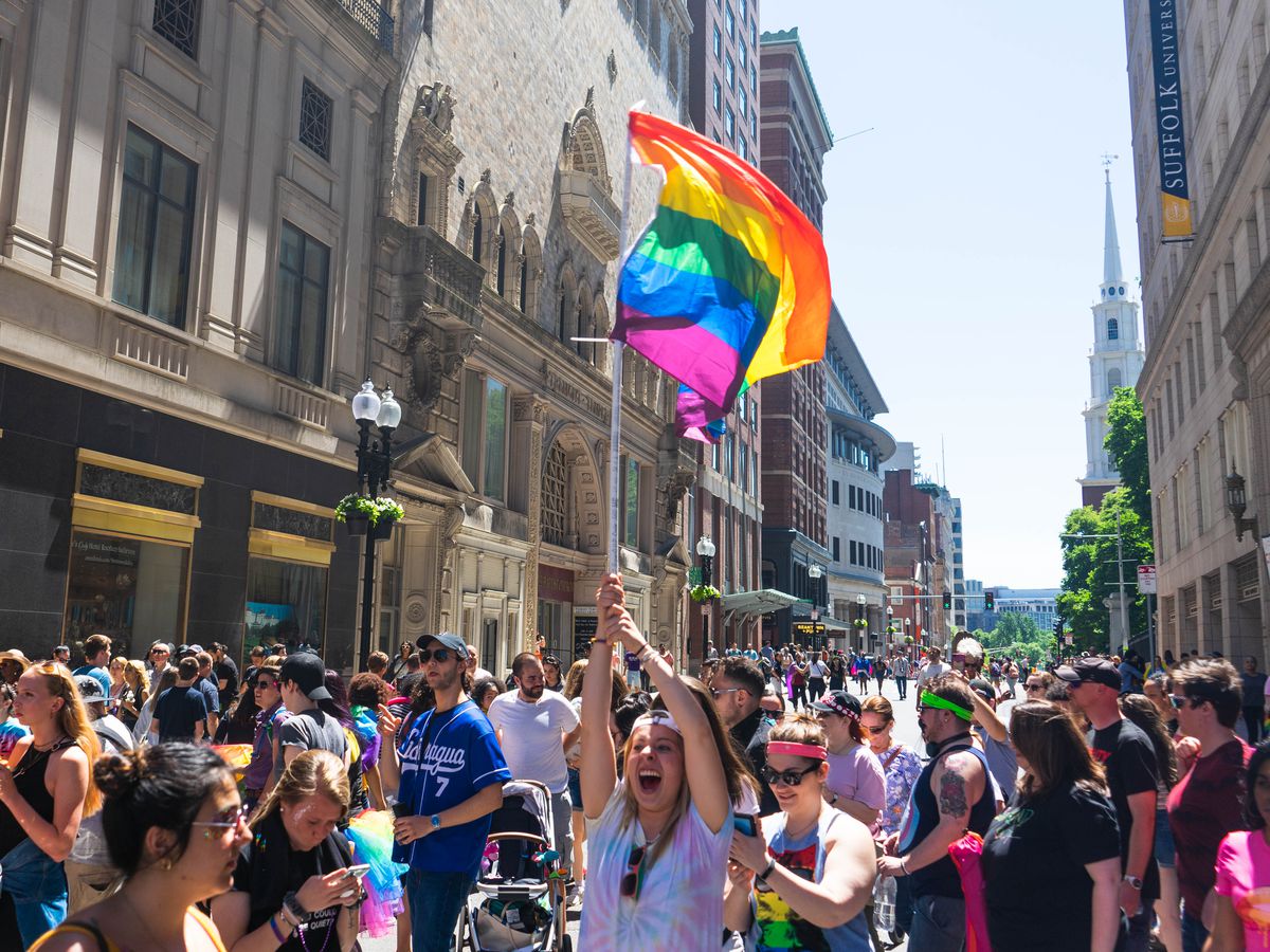 A woman in a crowd is shouting in glee and holding up a rainbow pride flag.
