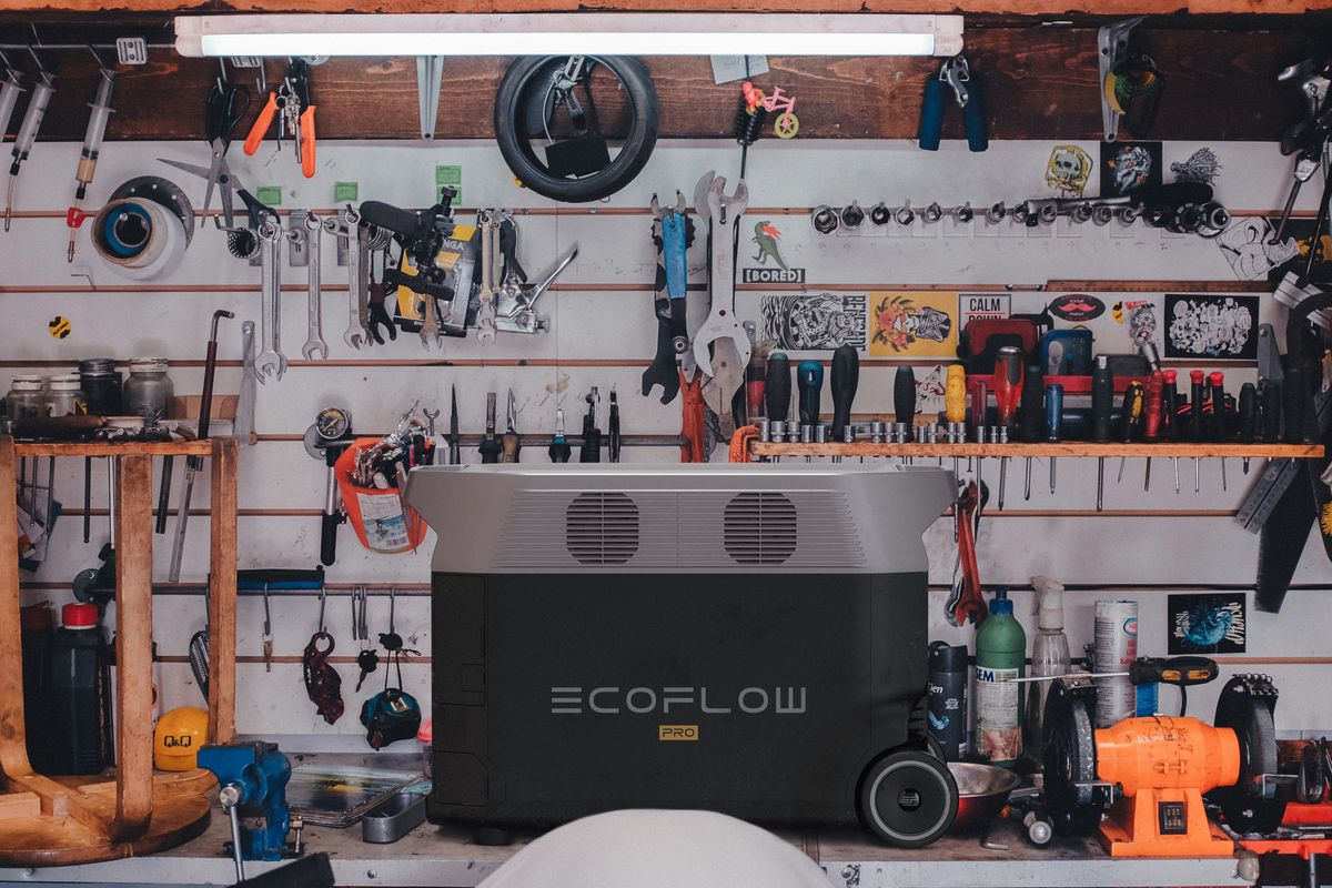 The EcoFlow DELTA Pro Power Station rechargeable battery sits in a tool shed, on a table in front of a wall full of tools such as wrenches and pliers. 
