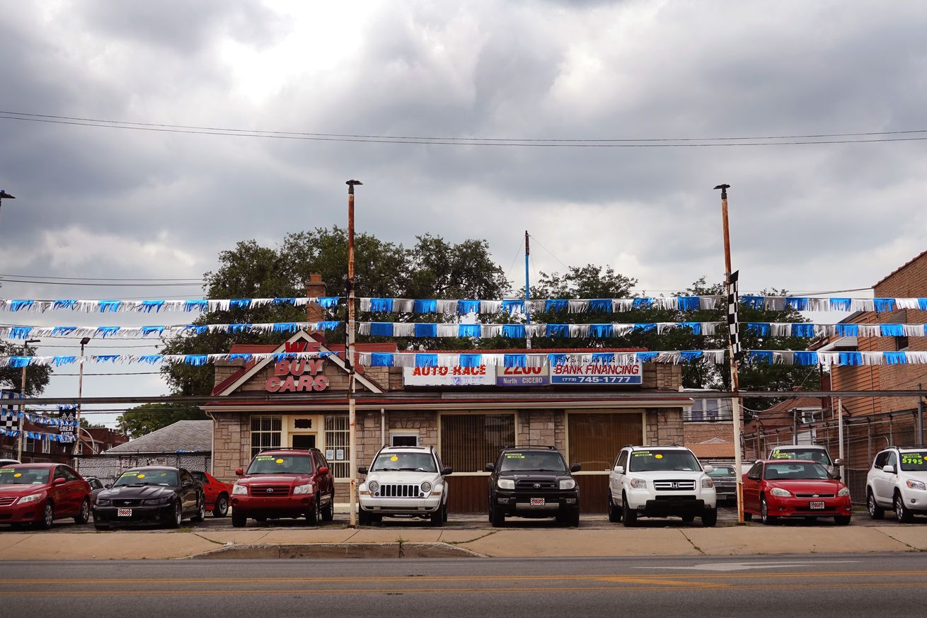 A picture of a car dealership showing the front of several cars in a lot, three blue and silver lines of streamers strung from poles above, and a small building behind them, on a cloudy day.