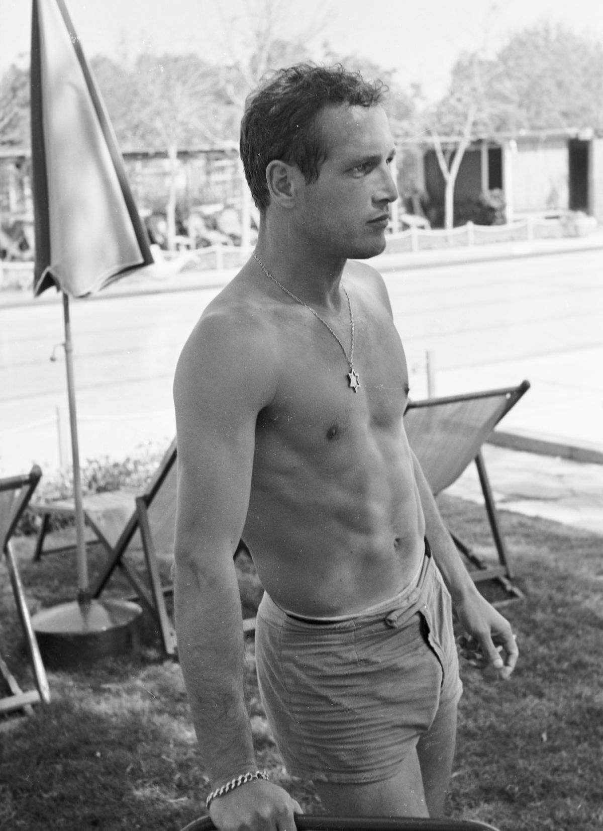 A shirtless Paul Newman in shorts smoking and leaning on a chair. He is wearing a chain necklace with a Star of David charm.