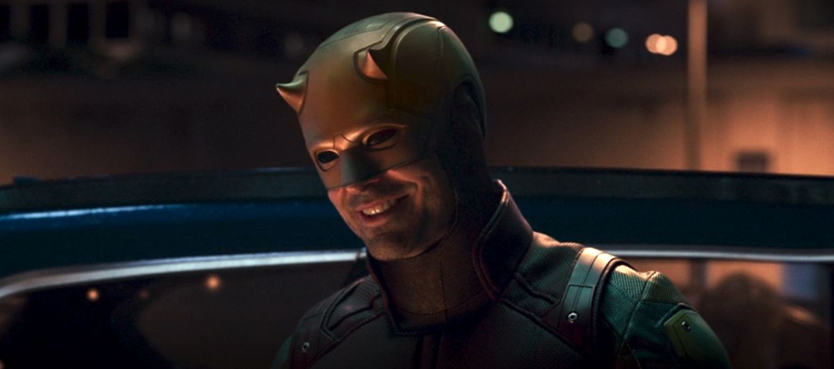 Charlie Cox as Daredevil in a yellow and red costume smiling in a parking lot at night