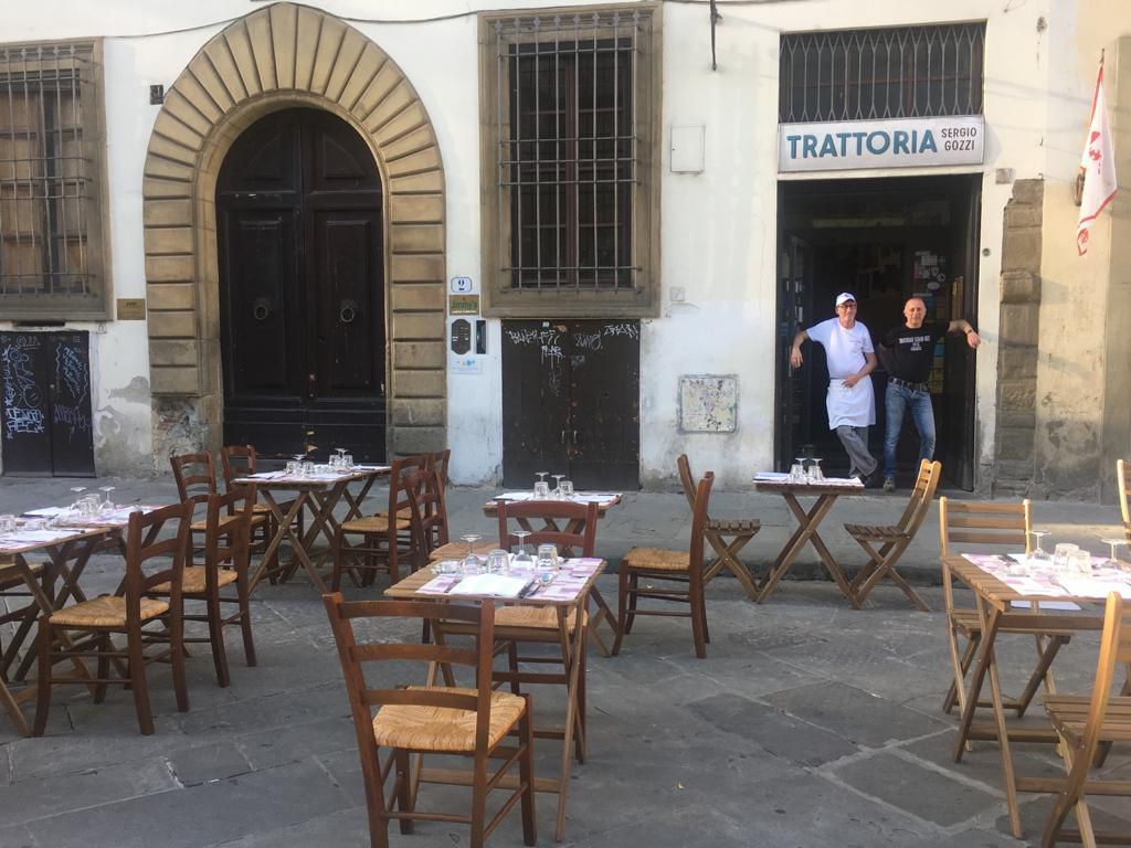 Street seating in front of a restaurant exterior, where two men lean in the doorway to the restaurant