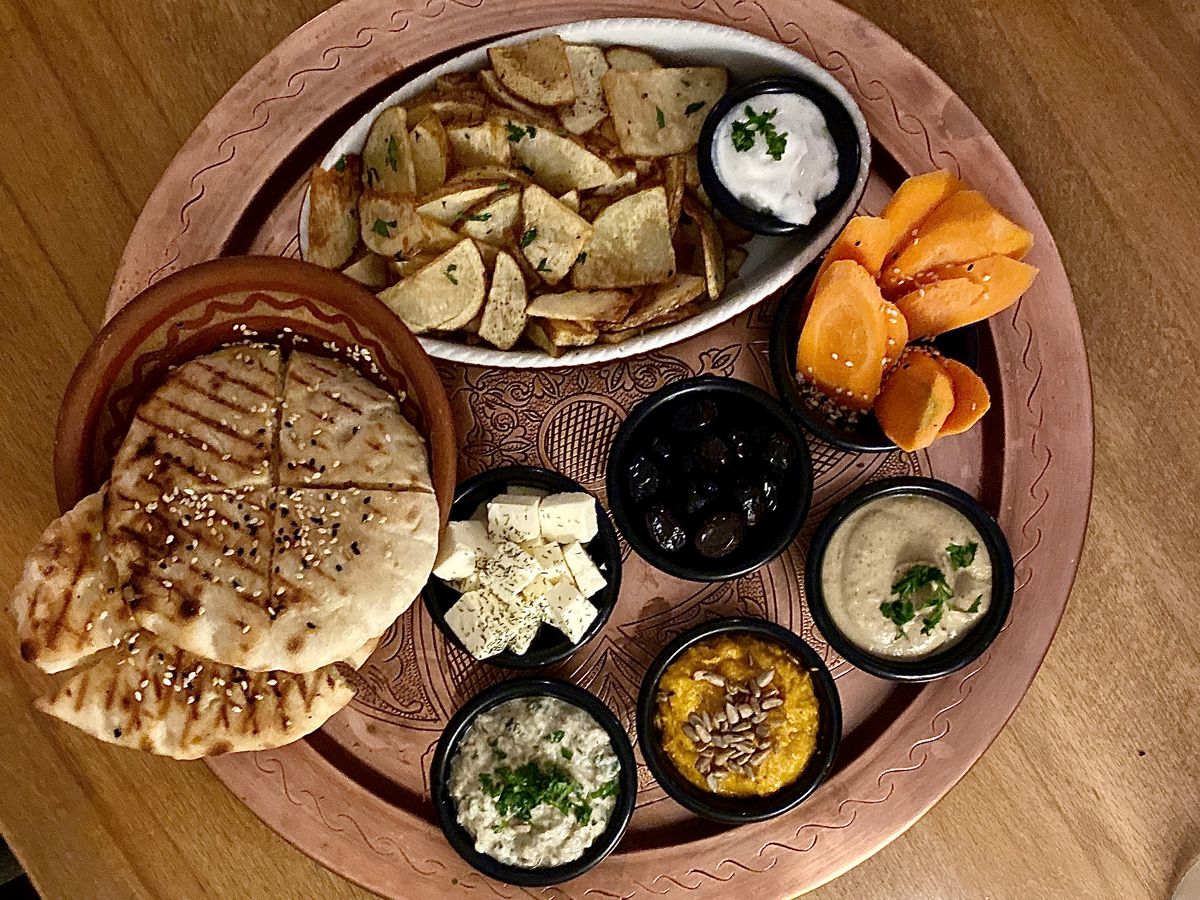 From above, a round wooden tray with large dishes of slices potatoes and pita, along with smaller cups of dips, carrots, olives, and cheese