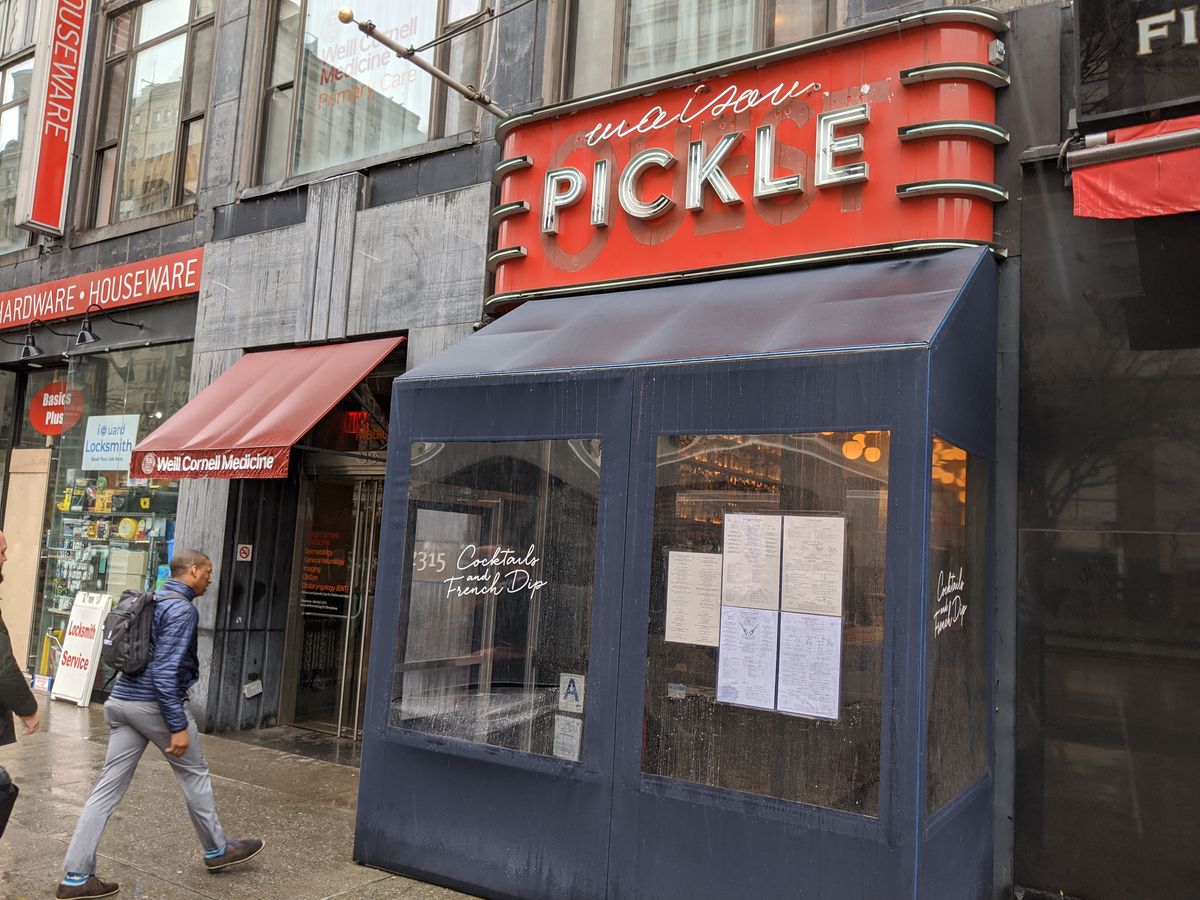 A person is walking into a gray restaurant with a neon sign that says Maison Pickle