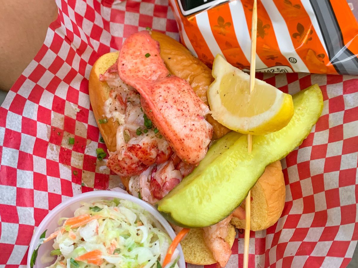 red and white checked paper beneath lobster roll, pickle, chips, and slaw