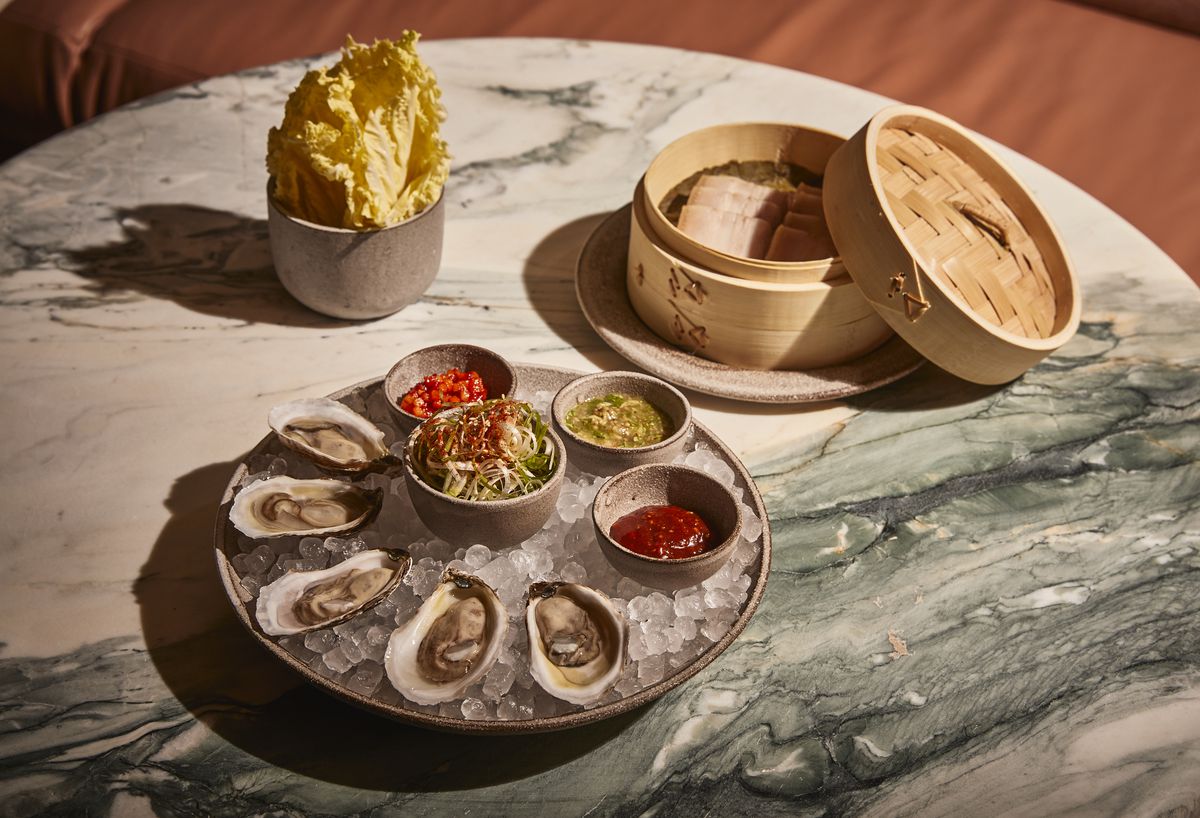 A platter of five oysters on ice besides colorful sauces and a bamboo steamer full of meat.