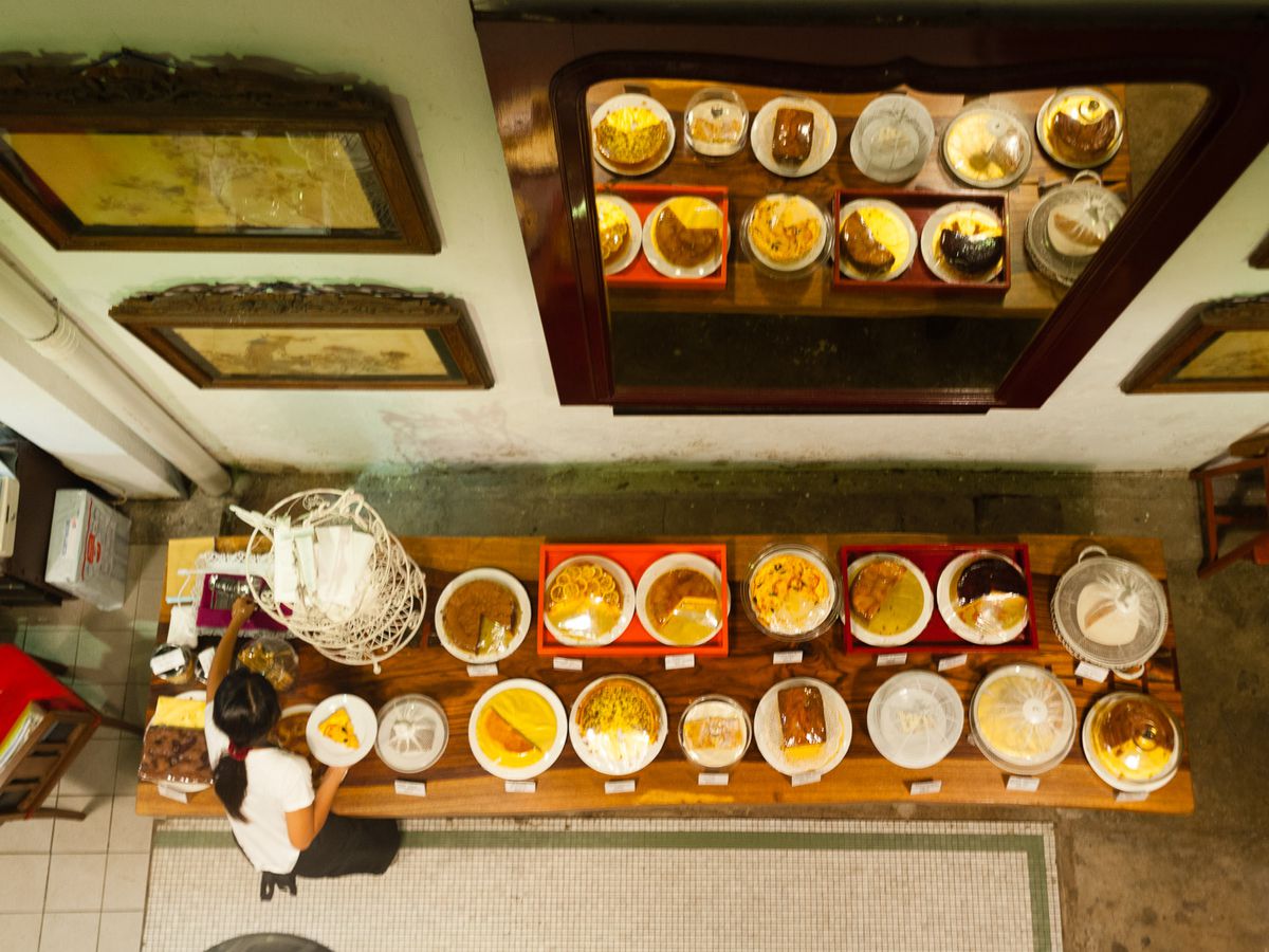 From above on a balcony perch, a server prepares a plate from a long wooden table of cakes and other desserts set against a wall decorated with a large mirror that reflects the offerings, as well as several obscured pieces of art.