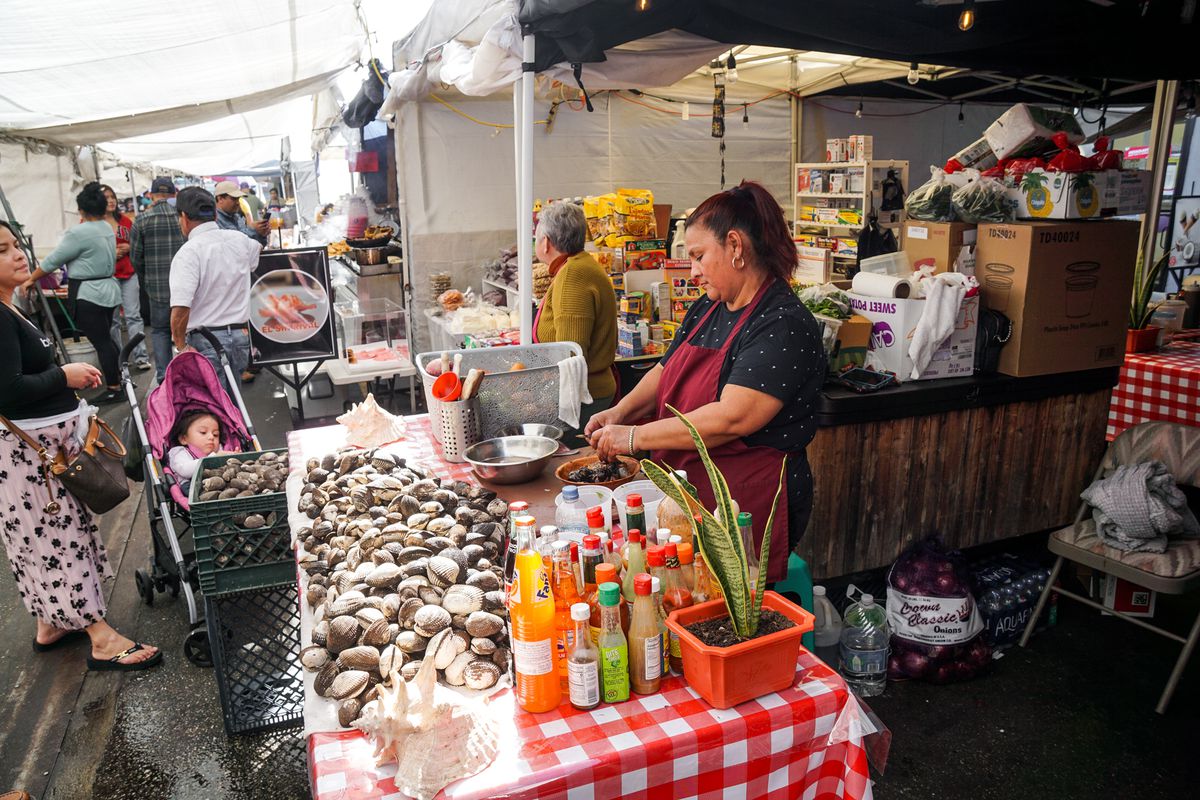 A vendor opens oysters and clams at an outdoor street food market in Los Angeles, with food situated on a checkered red table with customers passing by.