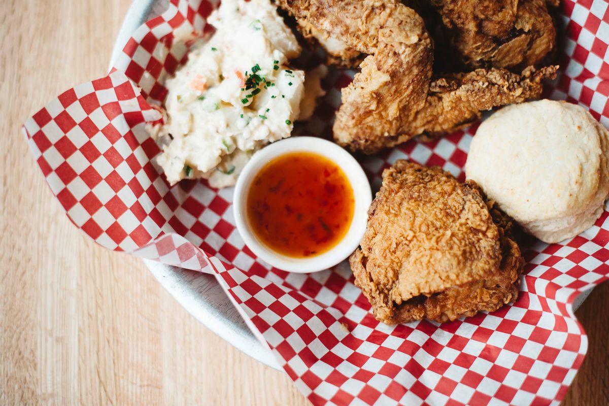 Overhead view of fried chicken, mashed potatoes, slaw, and dipping sauce on a red-and-white checkered paper on a white plate.