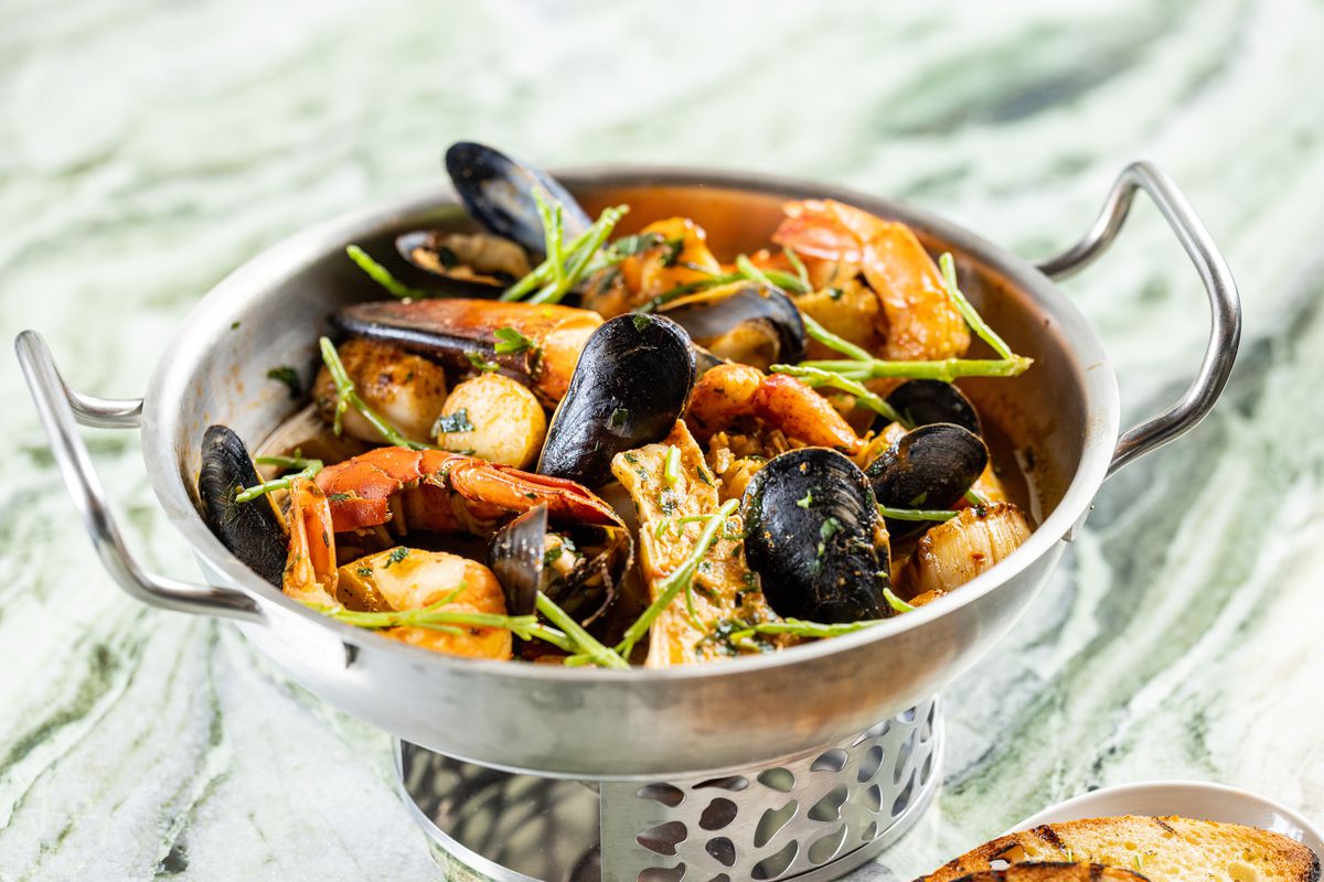 A basket of seafood in a silver boil basket with mussels, prawns, scallops, and more, served with toast. 