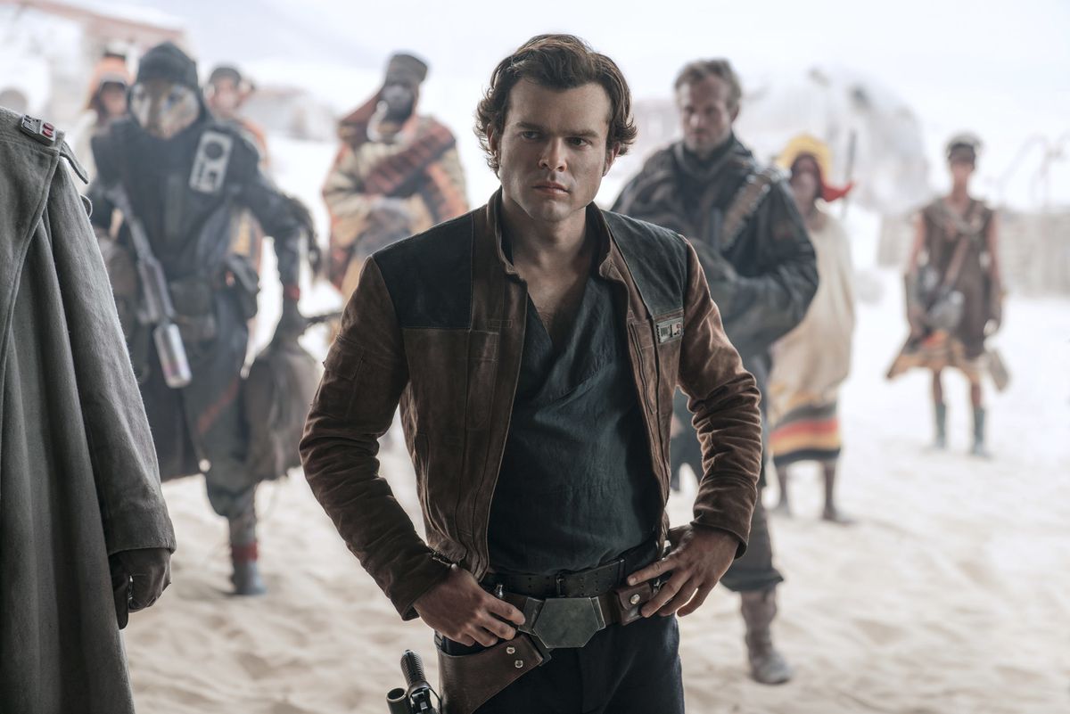 Alden Ehrenreich rests his hands on his hips while standing on sand, with people behind him, in Solo