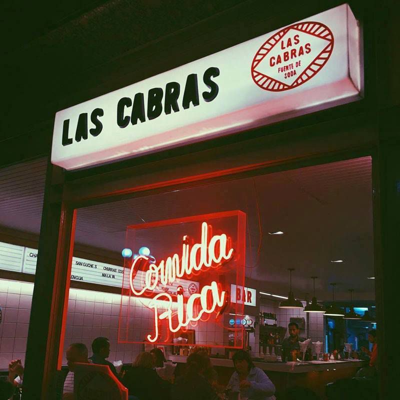 A window looking into a restaurant decorated with neon reading “comida rica” beneath a lit sign reading Las Cabras