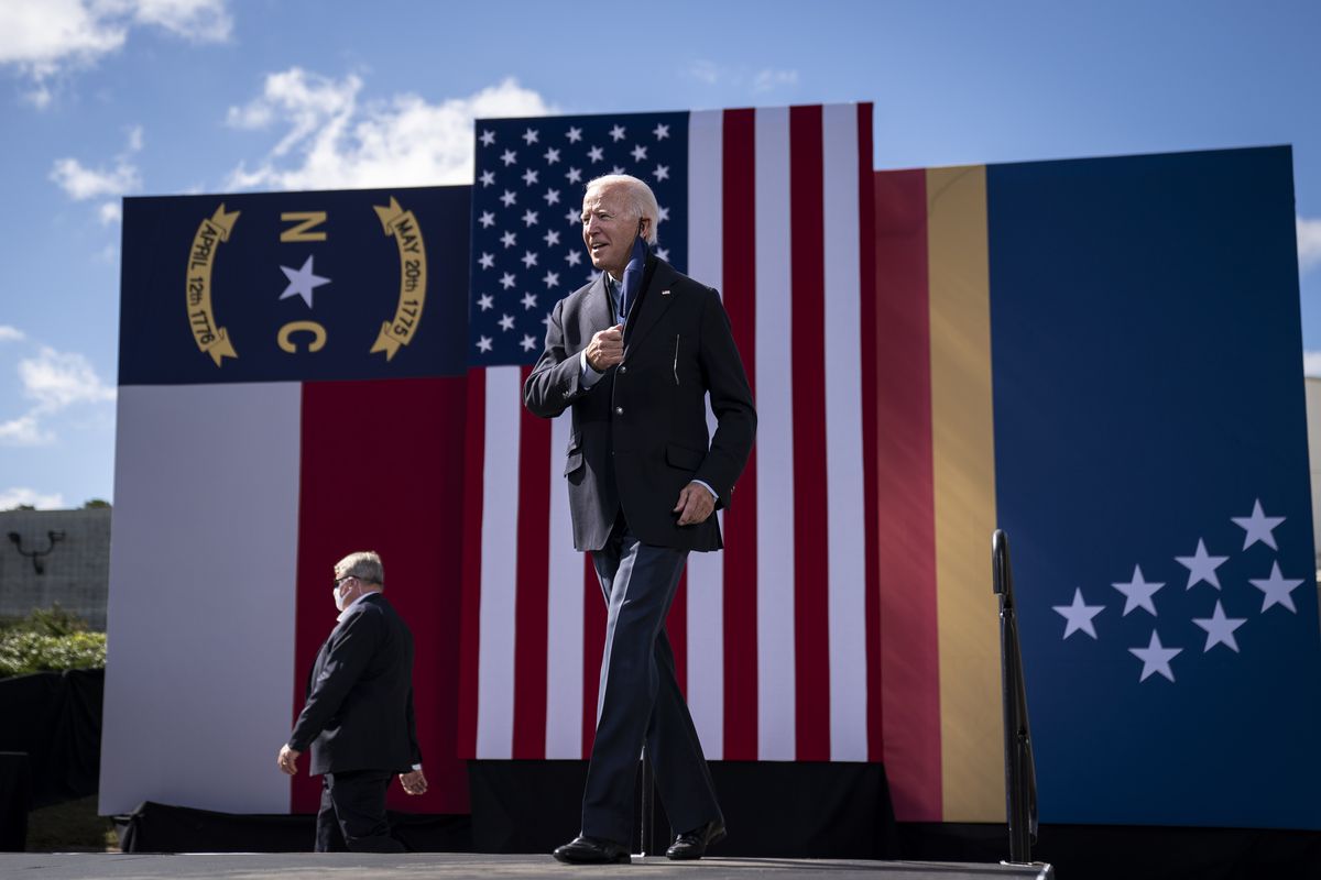 Biden pulls off a navy face mask as he mounts an outdoor podium, giant North Carolina, USA, and Durham flags behind him. The NC flag features red and white horizontal stripes, and the Durham flag a blue field with narrow red and yellow stripes at the bottom; 7 white stars are scattered across the blue field.