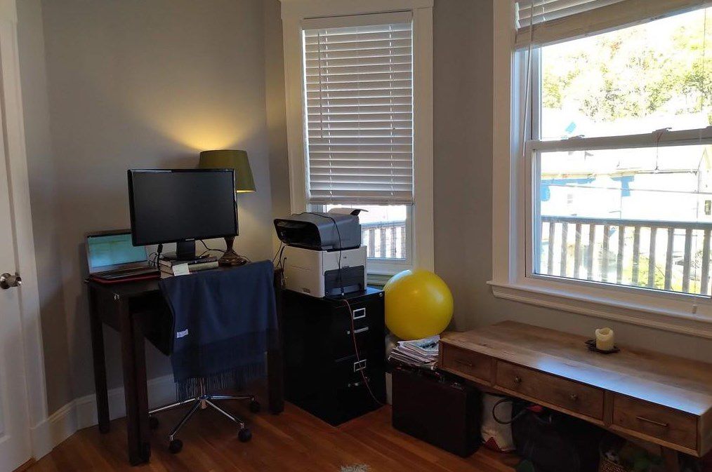 The corner of a small living room with windows, and the corner’s crowded with a filing cabinet and a desk with a computer.