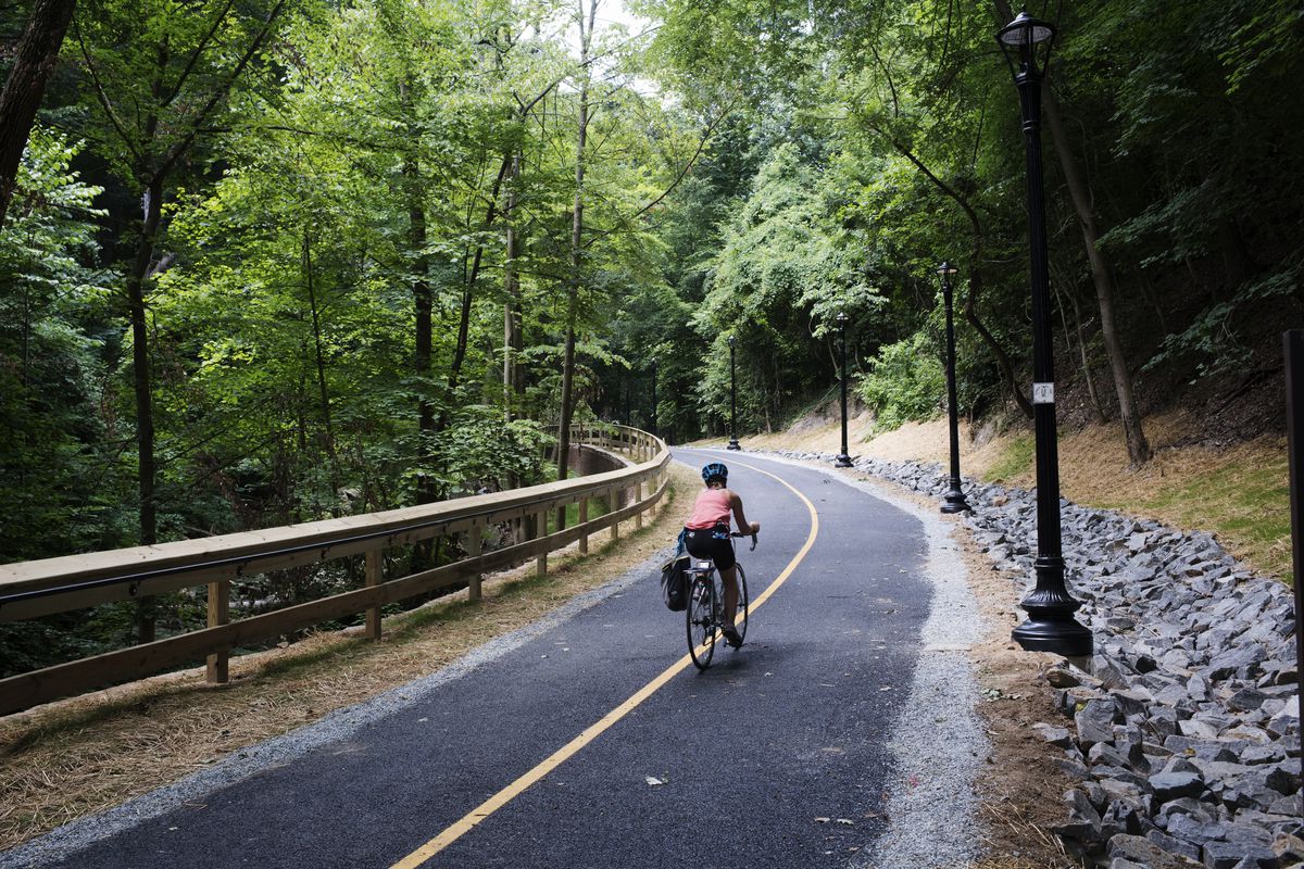 A cyclist uses a multiuse trail in the middle of a lush park. The trail is lined with streetlights.