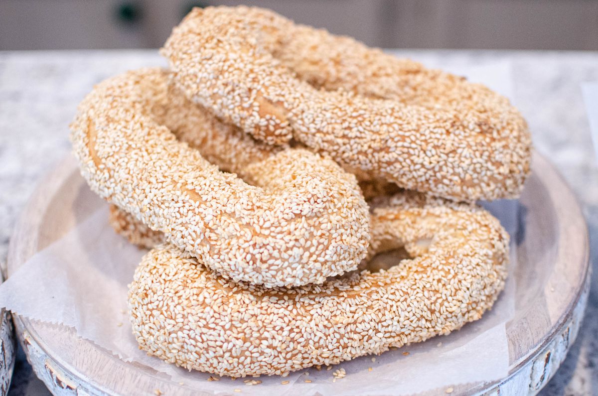 Four sesame seed-coated bread rings that look like oblong bagels are stacked on a plate that looks like a slice of a birch tree.