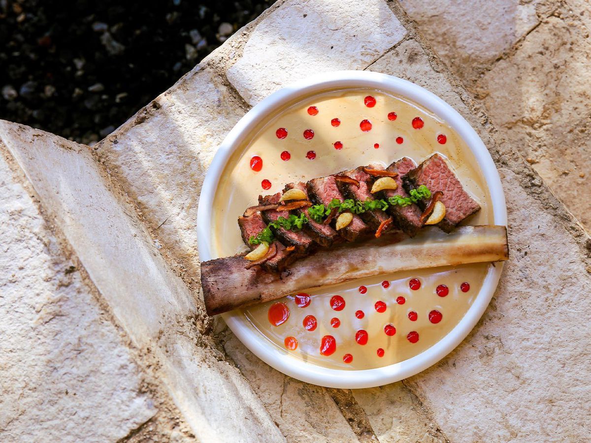 From above, a plate on stone steps, with a large short rib sliced with the bone, in a creamy sauce dotted with chile oil.
