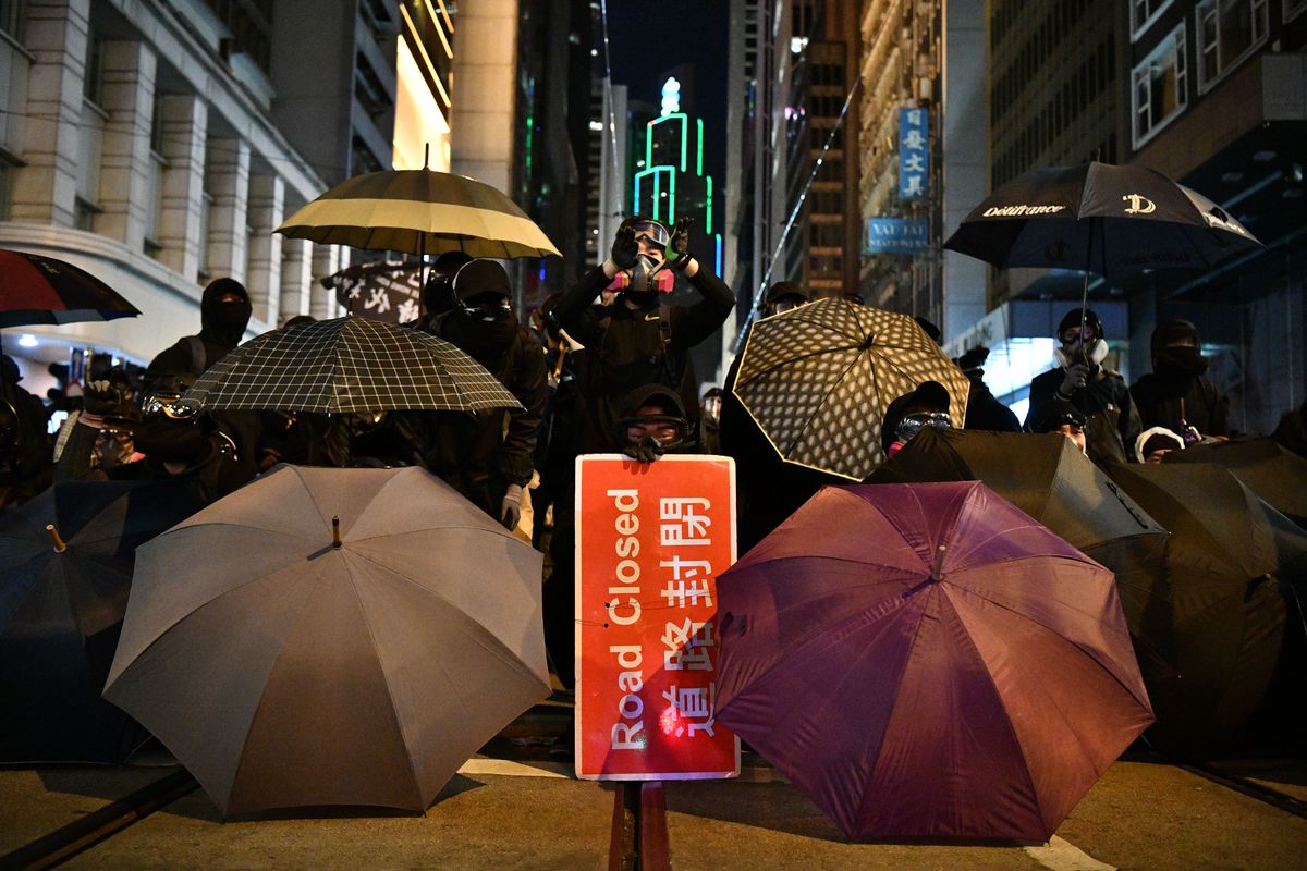 Black-clad protesters hold umbrellas and a sign saying “Road Closed” in front of building lit up for the night. 
