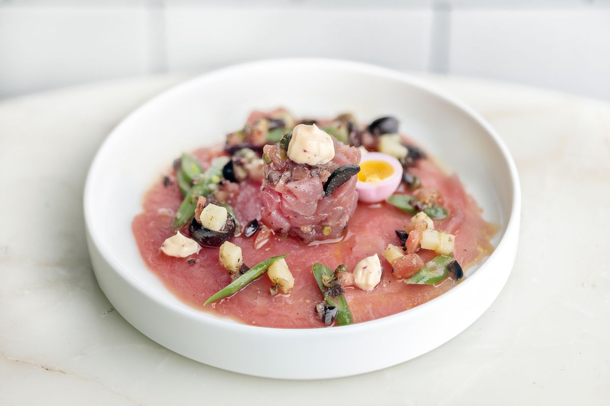 A pink yellowfin tuna crudo, garnished with a small egg and other Nicoise-inspired condiments, sits in a white bowl on a white marble table