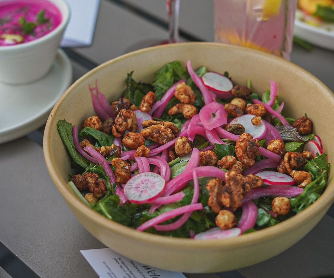 A bowl of greens topped with pickled onion, radish, and chickpeas, on an outdoor table beside a colorful cocktail