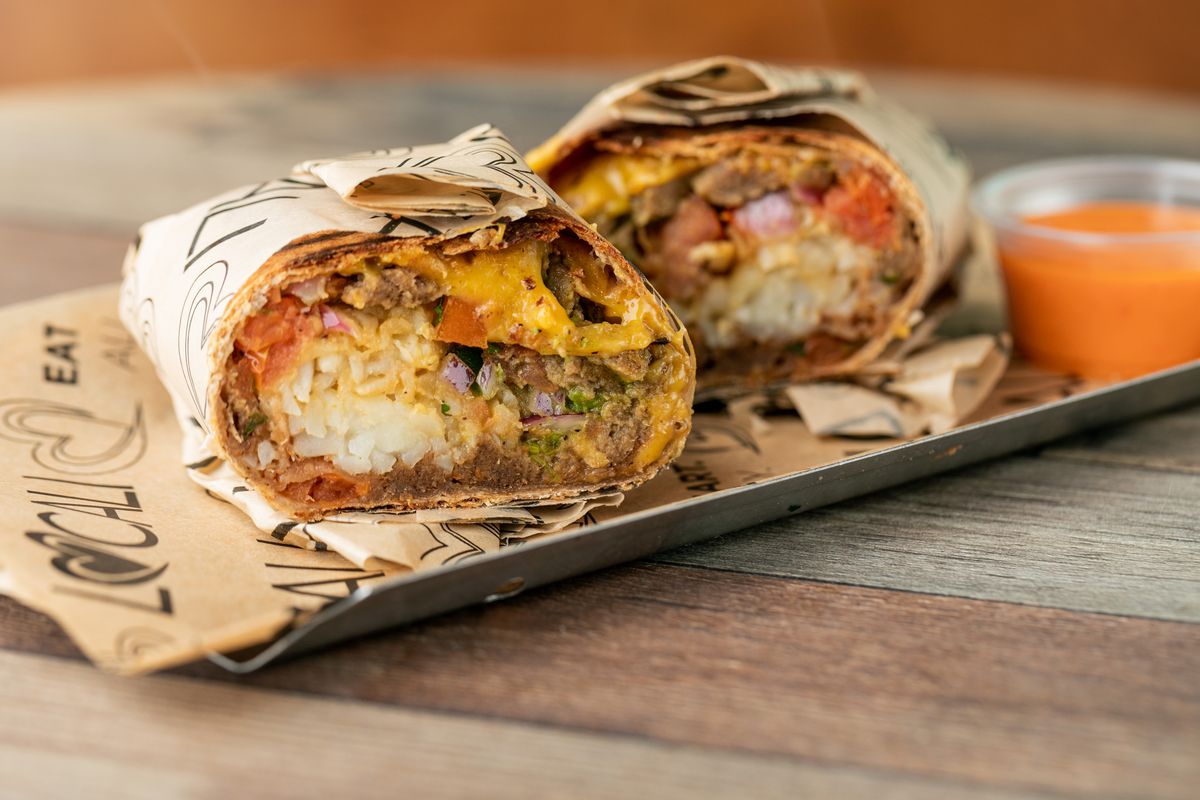 A burrito cut in half, showing rice, vegan meat, and cheese inside, next to a cup of orange sauce on a tray.