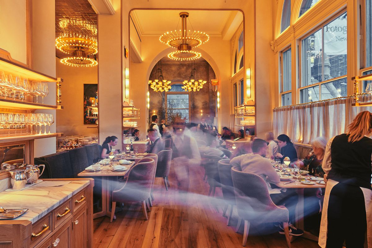A tall-ceilinged dining room with golden tones and big chandeliers, with waiters zooming by.