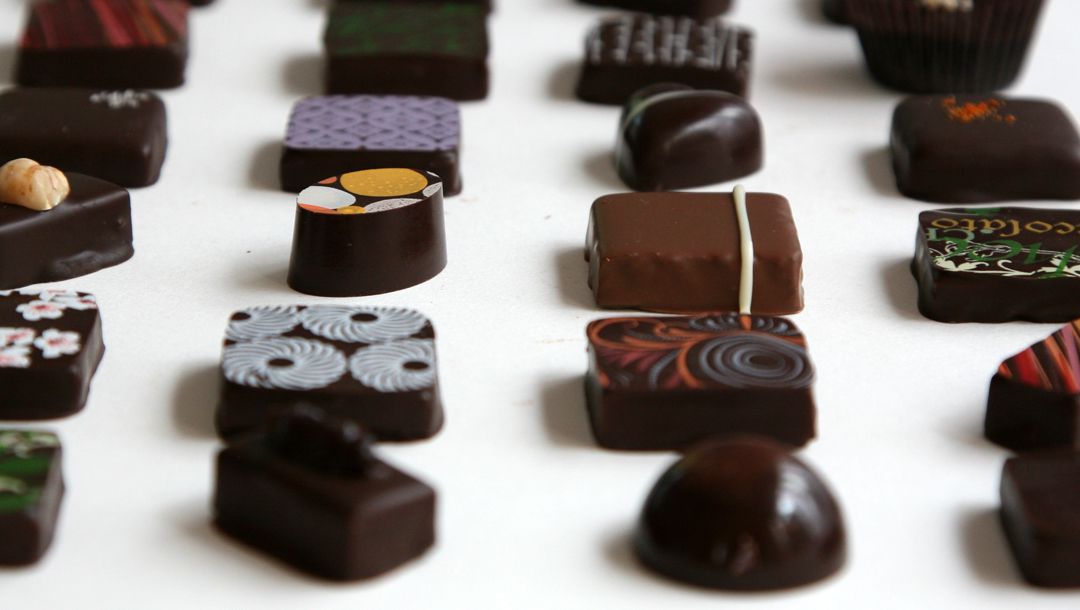 Several rows of chocolate bonbons, with a variety of shapes, sizes, and decorations, are lined up on a white background.