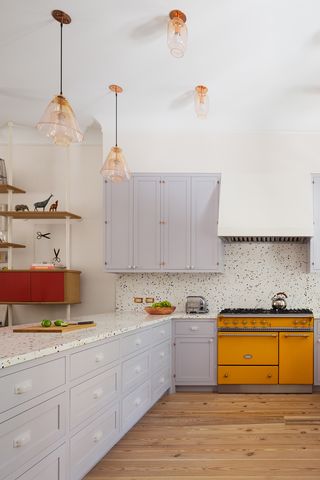 kuchyň s přírodními dřevěnými podlahami, levandulovými skříněmi, žlutým sortimentem a teracovými deskami a backsplash. 