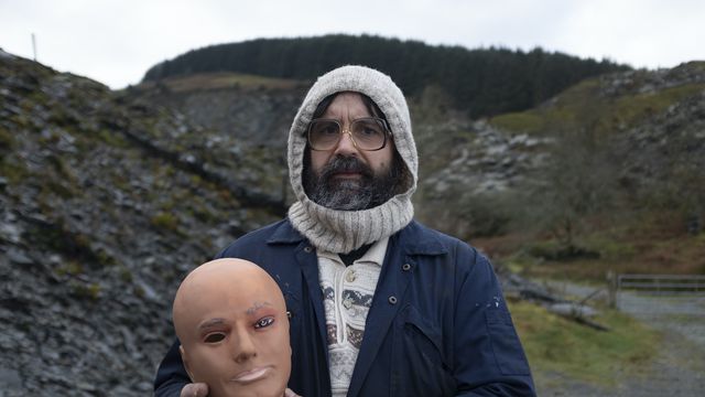 Brian (David Earl) stands among stone piles, holding a mannequin head, in Brian and Charles