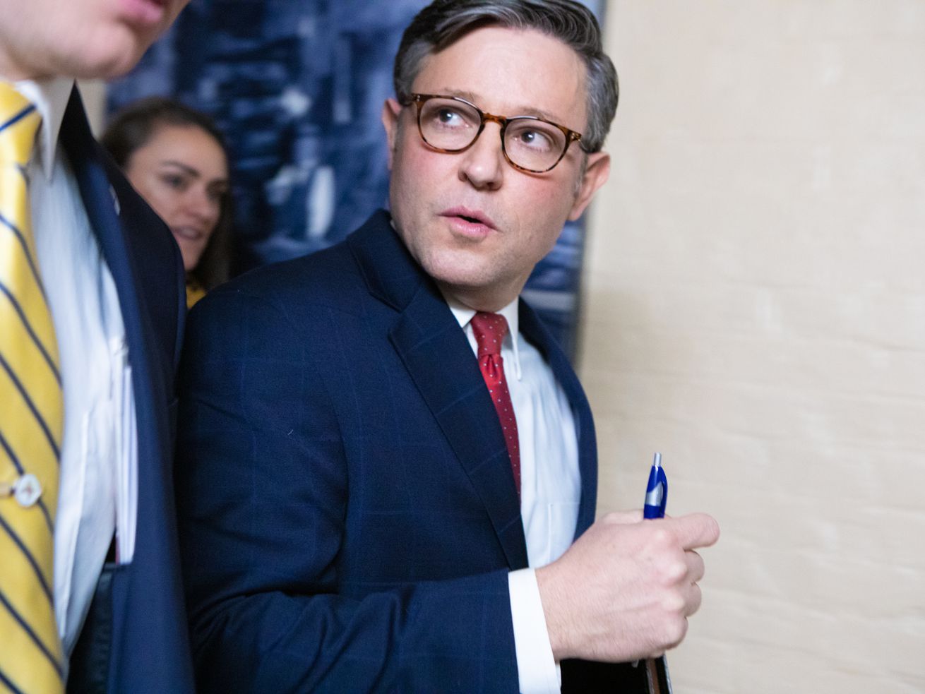 Mike Johnson pictured wearing a navy suit jacket, red tie, and glasses. He appears to be holding a blue pen in his hand.