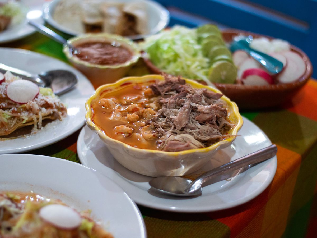 A decorative bowl filled with pozole, including mounds of meet sticking out of the broth, alongside other dishes