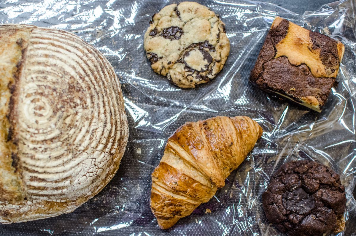 Overhead view of a round loaf of bread, a chocolate chip cookie, a croissant, a marble brownie, and a fudge cookie on a piece of plastic on a dark table