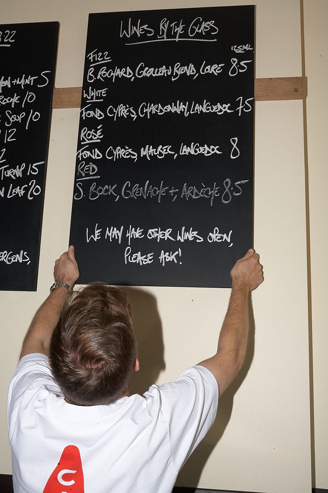 A man in a white t-shirt puts up a blackboard with wines by the glass written in white chalk.