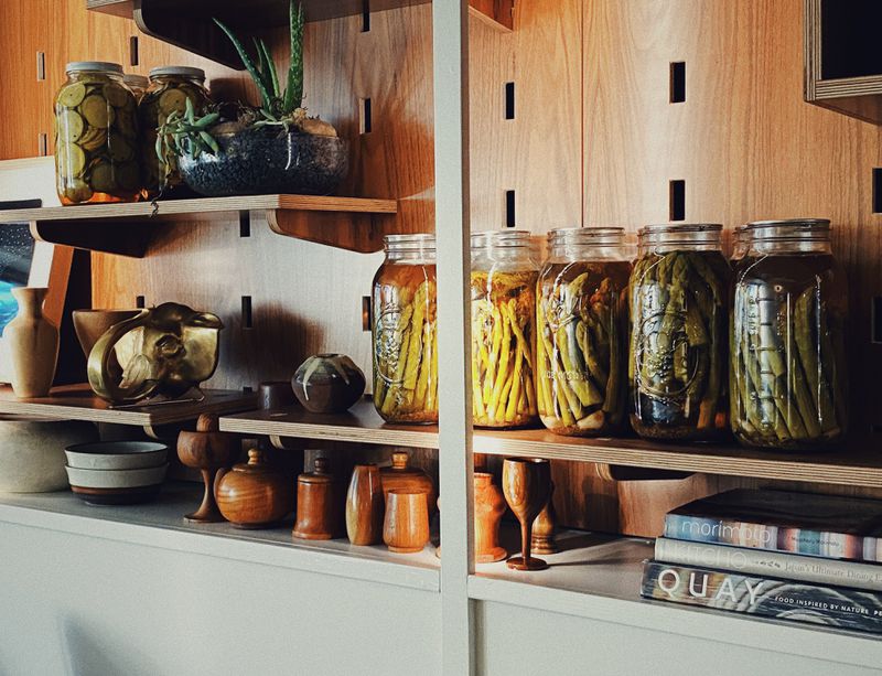 Jars of brightly colored vegetable pickles on shelves beside wooden serveware, art, and cookbooks
