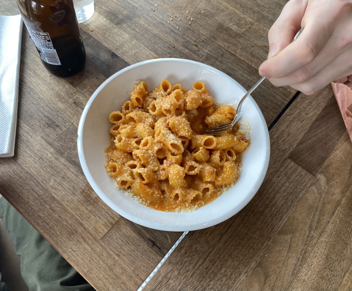 Een hand bungelt een metalen vork in een kom met oranje pastanoedels bedekt met kruimelige Parmezaanse kaas.