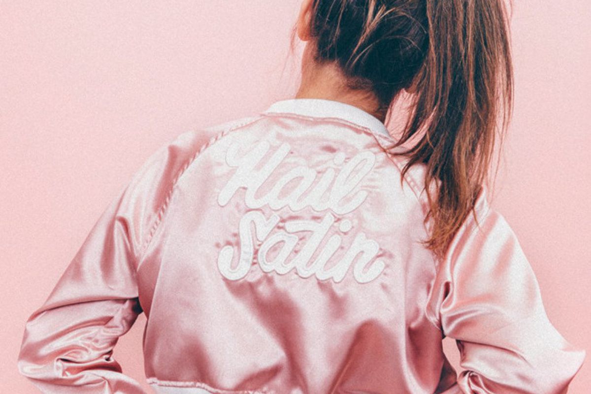 A girl with a ponytail wearing a pink silk bomber jacket that says “Hail Satin”
