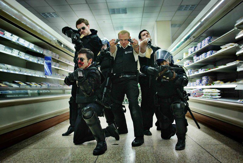 Simon Pegg as PC Nicholas Angel stands with his fellow officers in a well-lit shopping aisle, hold twin pistols akimbo style