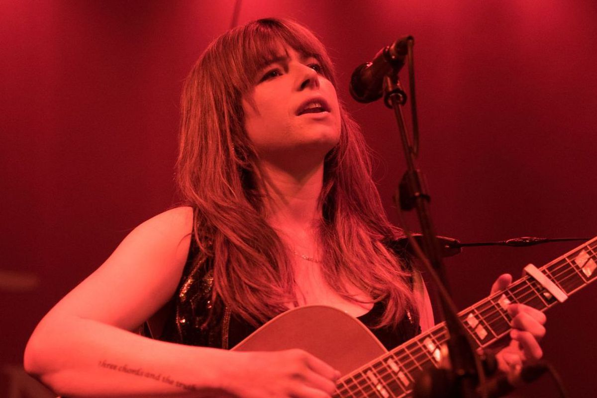 A young woman plays guitar and sings, bathed in a red light.