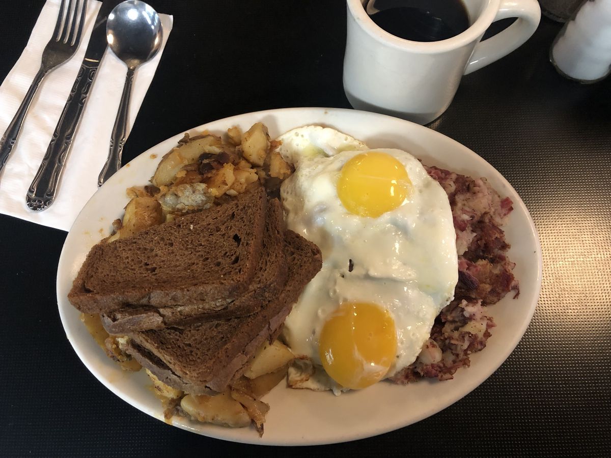 A white oval plate filled to the brim with potatoes and hash and two sunny-side up eggs and four half-pieces of rye toast set on top.