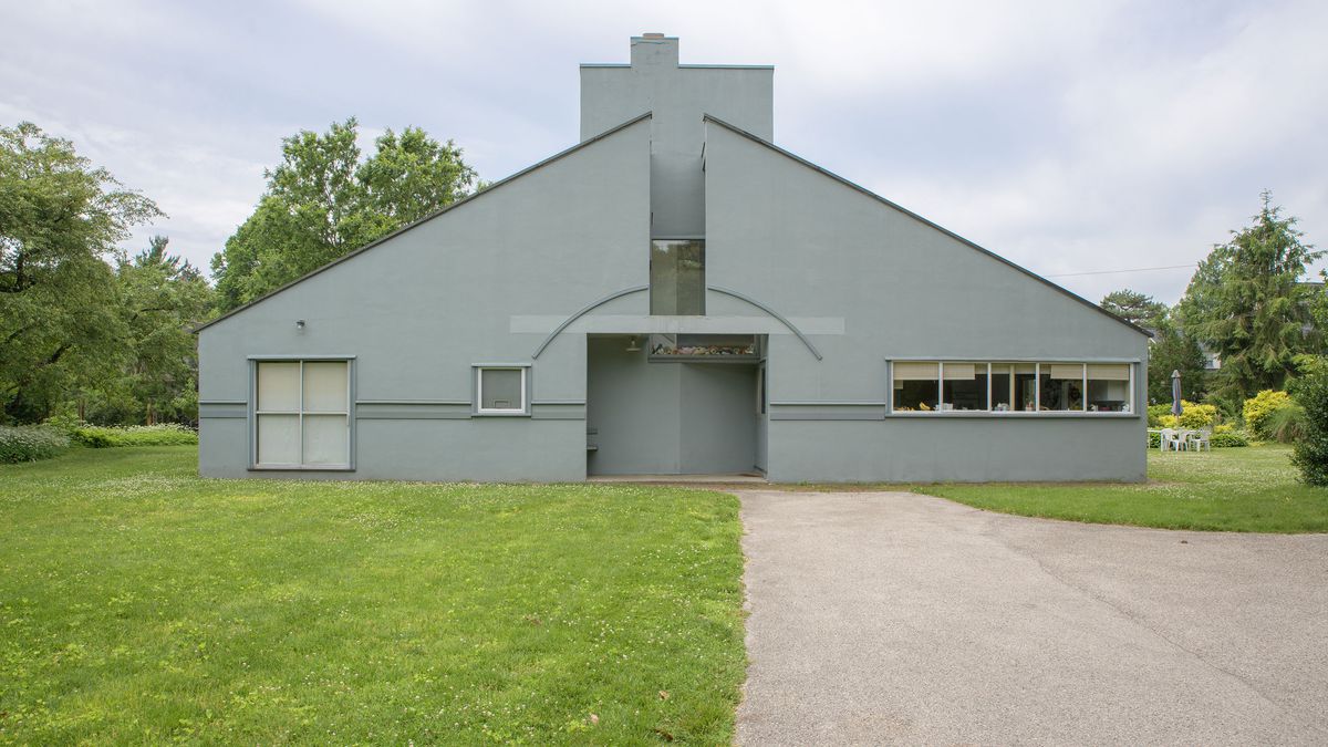 The Vanna Venturi house in Philadelphia. The exterior of the house is light blue with a sloped roof and large entrance. There is a path leading up to the entrance. There is a large green grass lawn adjacent to the path in front of the house.