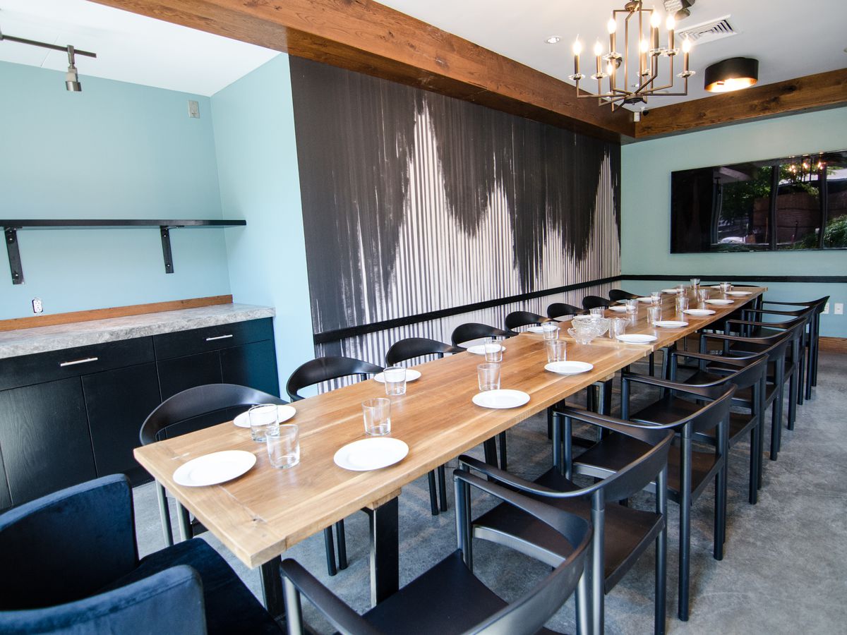 A long wooden table with about 18 chairs stretches through a private dining room at a restaurant. The walls are light blue, except for one portion, which is covered with a black-and-white-striped wallpaper. 