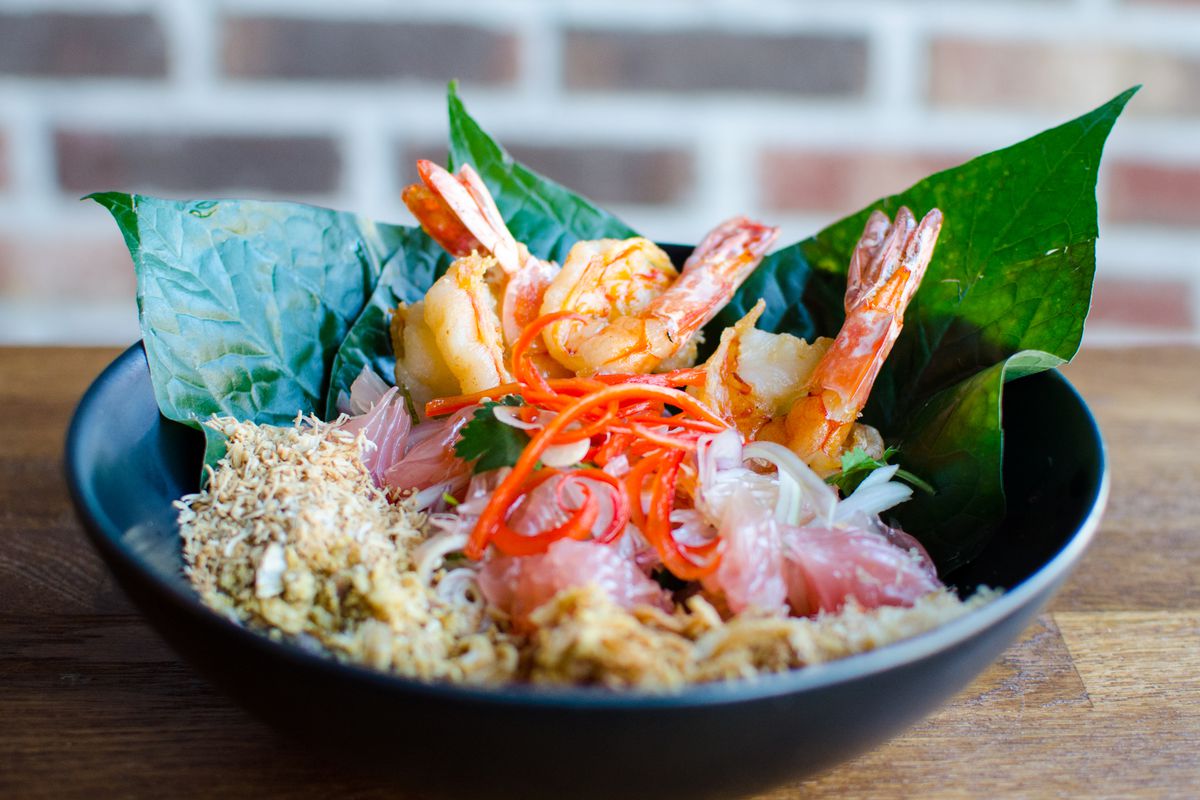 A Thai pomelo salad with shrimp, betel leaves, thinly sliced red chile, and a variety of crispy condiments sits in a black bowl on a wooden table in front of a brick background.