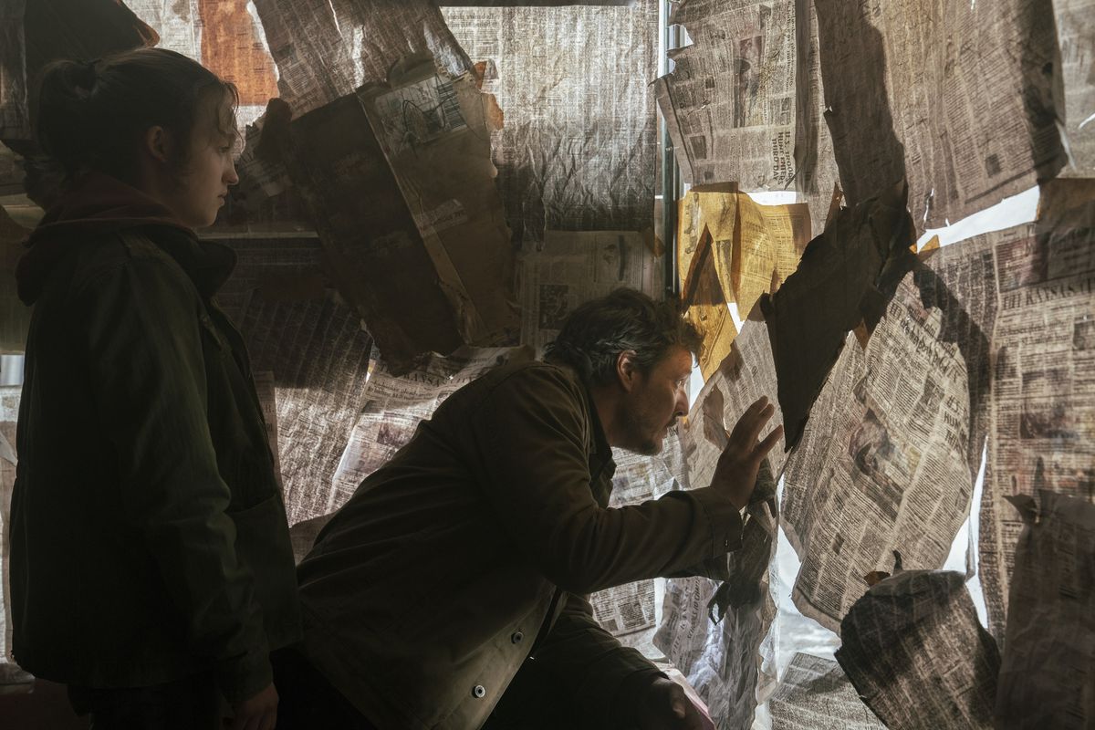 Joel (Pedro Pascal) looking out a window between newspapers while Ellie (Bella Ramsey) looks on