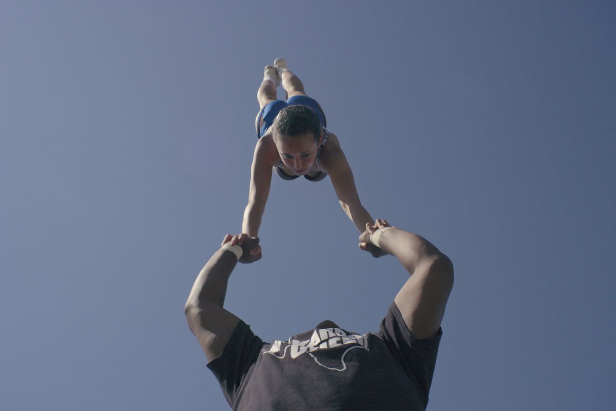 A male cheerleader holds a young woman in a handstand over his head.