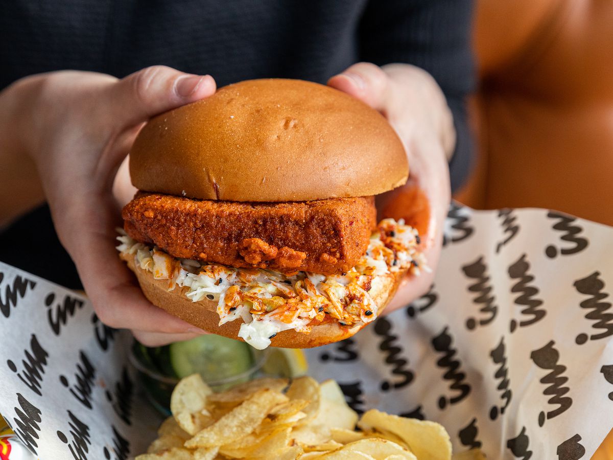 Hands hold a karaage hot tofu sandwich over a basket filled with potato chips at Ima.