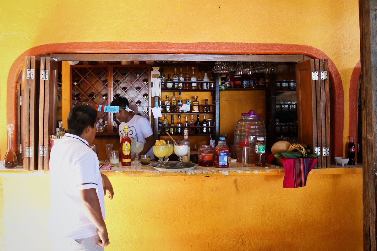 Un mesero se para en la apertura de un bar amarillo donde un hombre coloca cócteles en una bandeja. 