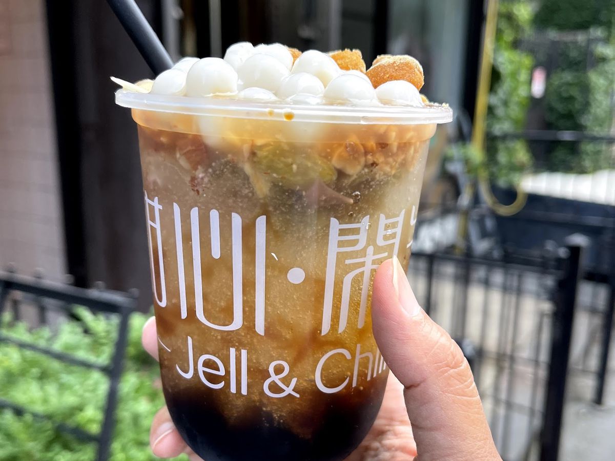 A hand holding a clear plastic cup of bing fen with haw flakes and mochi toppings.
