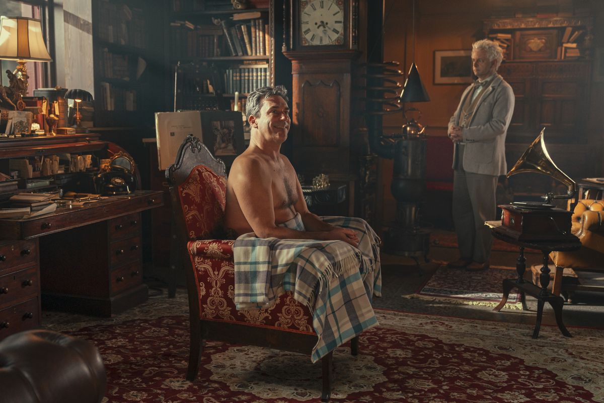 Jon Hamm as Gabriel, sitting completely naked in an arm chair in the middle of a bookshop, a tartan blanket draped over his lap. He is grinning widely. In the distance is Aziraphale, a man dressed in white, clasping his hands together and looking quite worried.